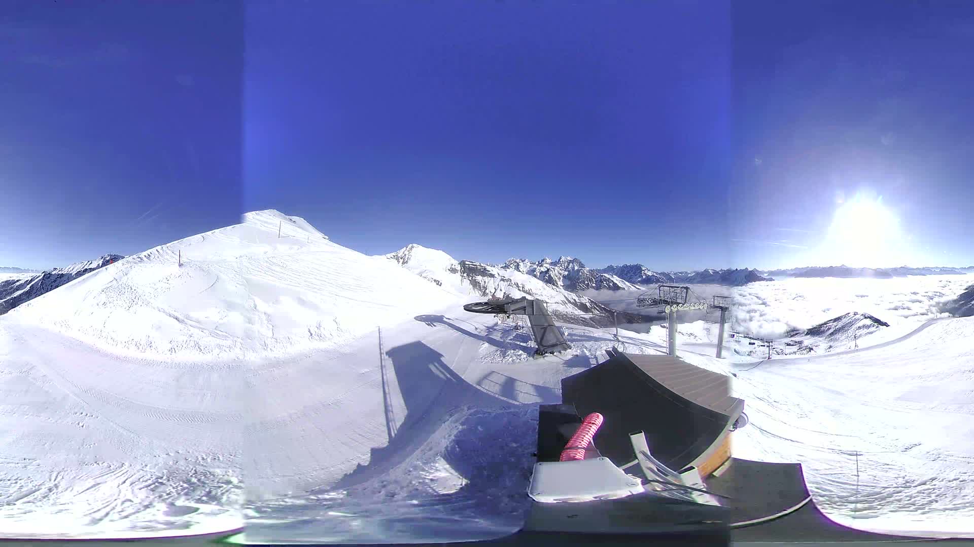 A sunny, snow-covered mountaintop with a ski lift and a view of a valley filled with clouds.