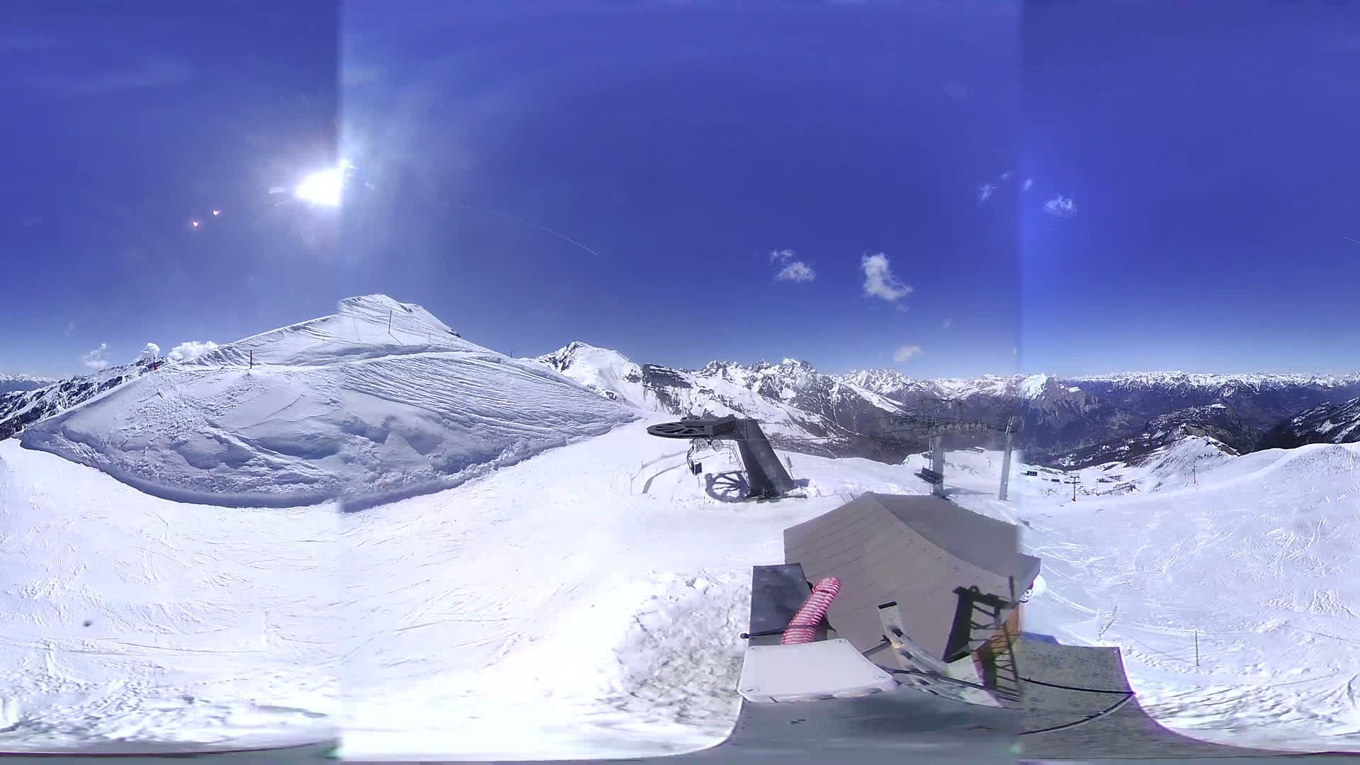 A sunny, snow-covered mountaintop ski resort scene features a ski lift, a small building, and expansive views of snow-capped mountains under a clear blue sky.