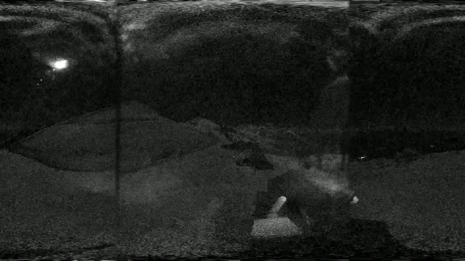 A nighttime, grainy image shows a person sitting on the ground in a dark, possibly sandy area under a cloudy sky illuminated by a distant light source.