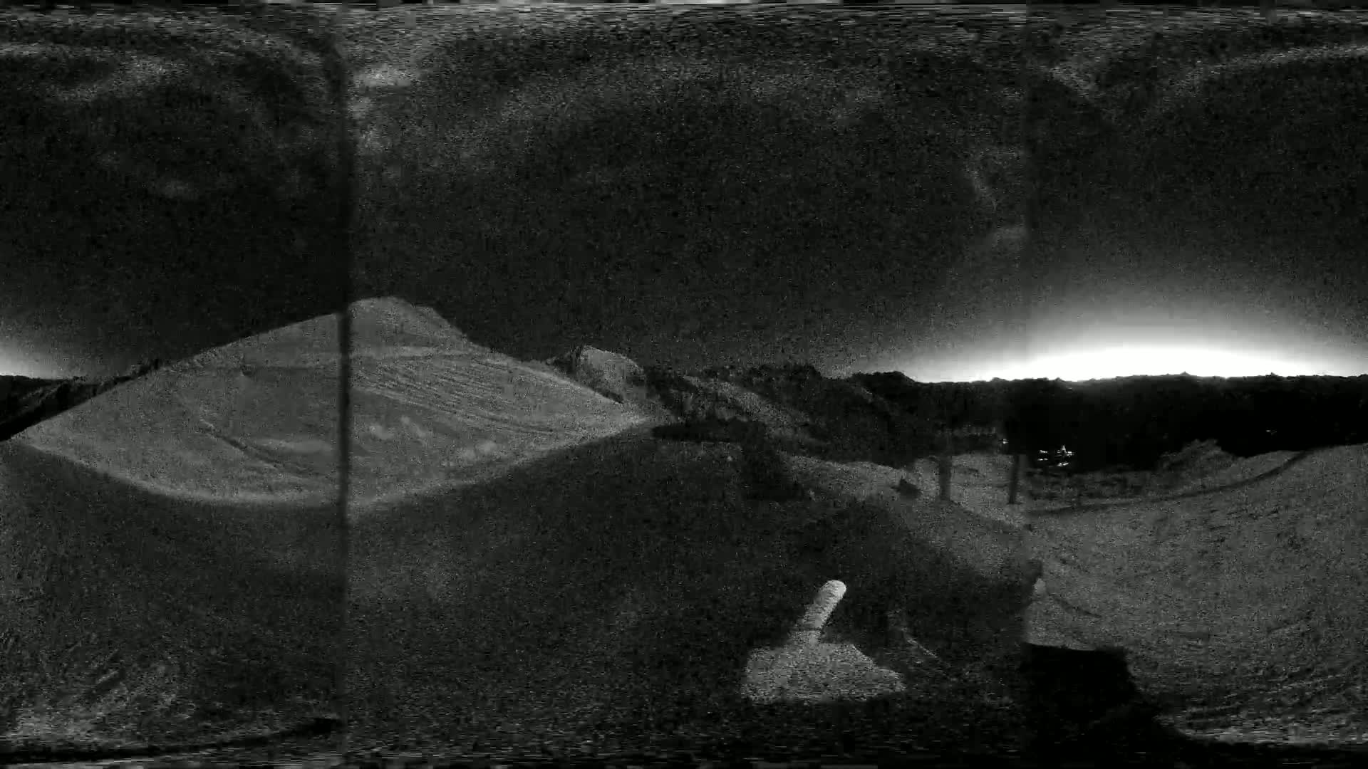 A grainy, black-and-white, panoramic view shows a dark, sandy landscape under a bright, possibly sunlit, horizon.