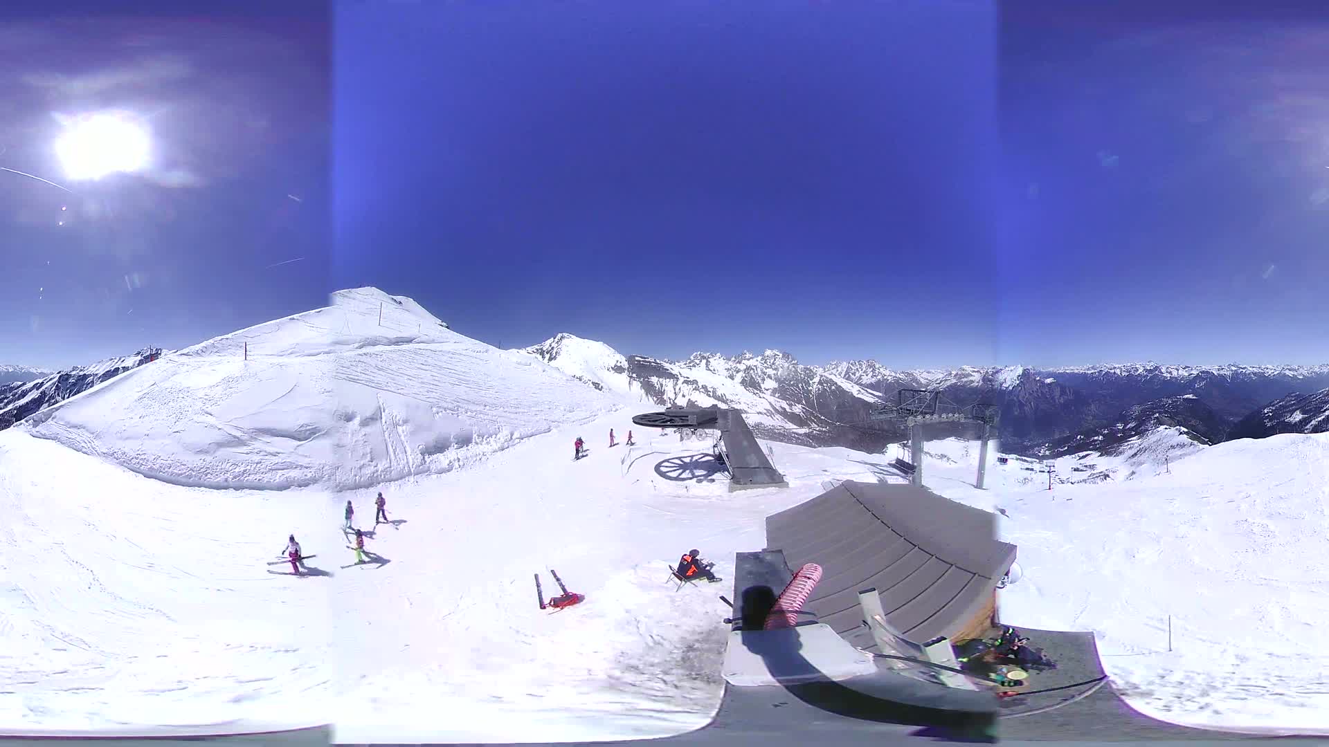 A sunny day on a snow-covered mountaintop shows skiers and snowboarders on a slope near a ski lift and mountaintop building, with a mountain range in the background.