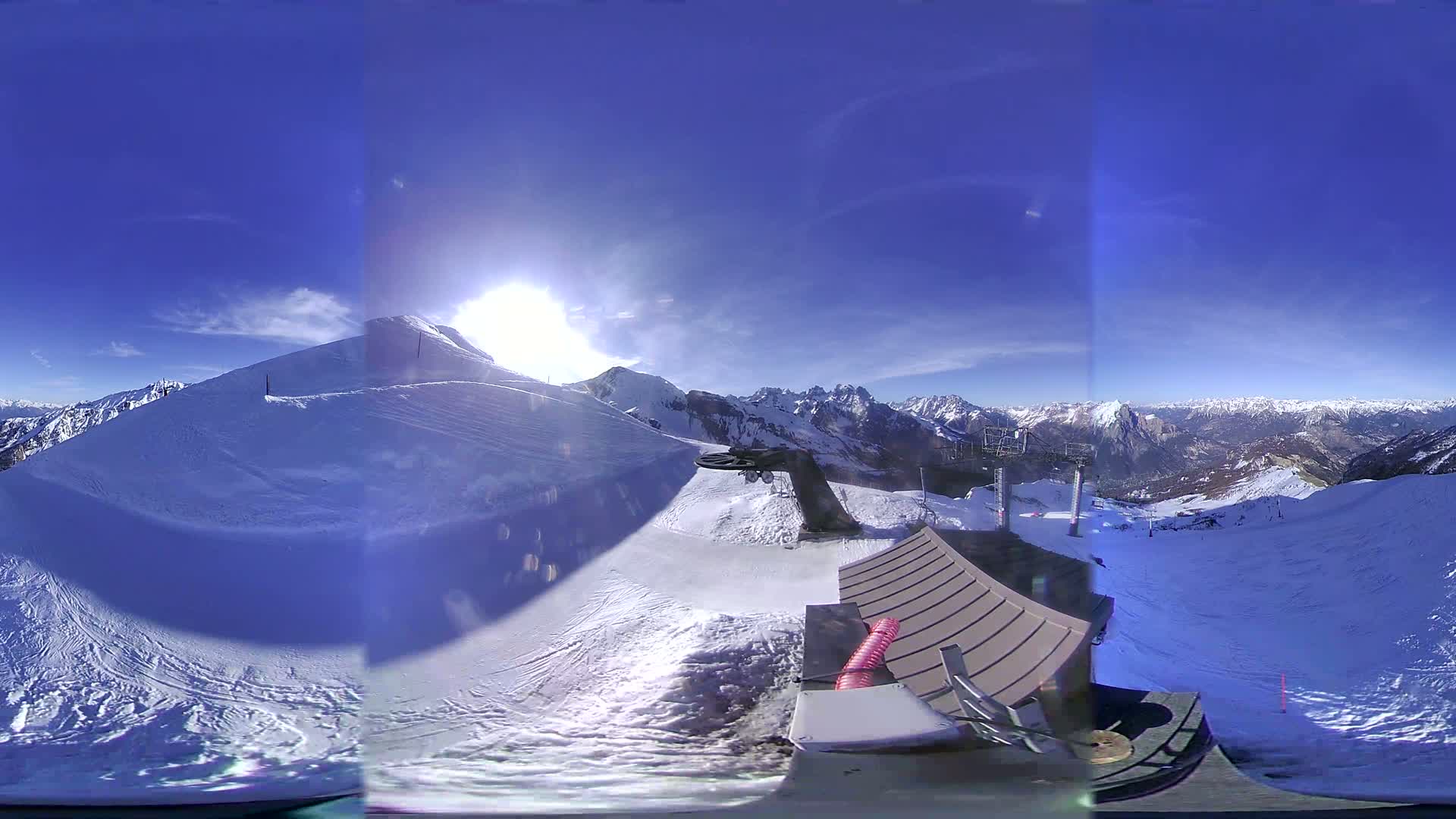A sunny, clear day on a snow-covered mountaintop reveals a ski lift and expansive views of a mountainous landscape.