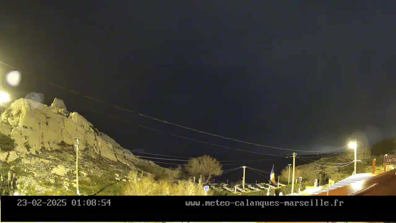 A nighttime view of a rocky hillside under a dark, clear sky, with some sparse vegetation and a building partially visible in the lower right corner.
