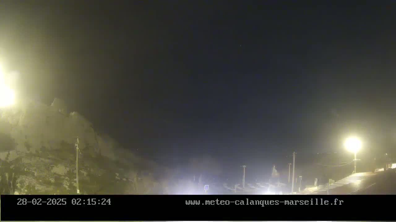 A nighttime view shows a hazy, dimly lit coastal area with a rocky cliff face to the left and a road with several lights in the foreground.