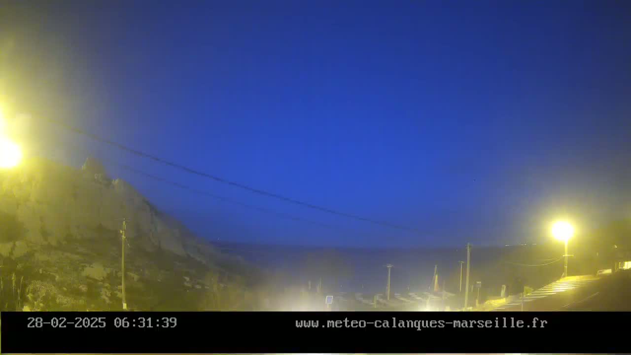 A hazy, dark-blue twilight sky illuminates a rocky hillside and a distant body of water, with streetlights casting a yellowish glow on the scene.