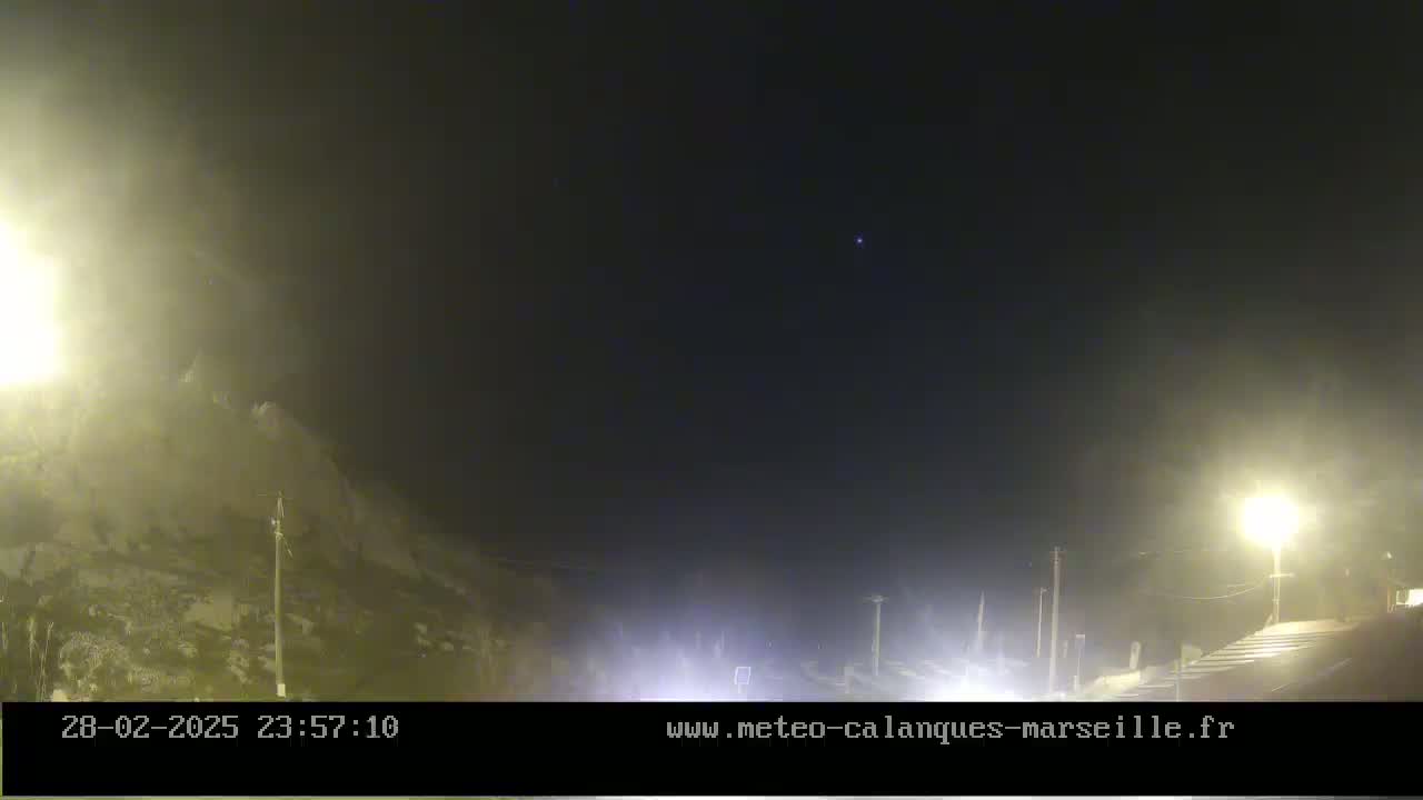 A nighttime, foggy scene shows a road with lights and a rocky hillside under a dark sky.