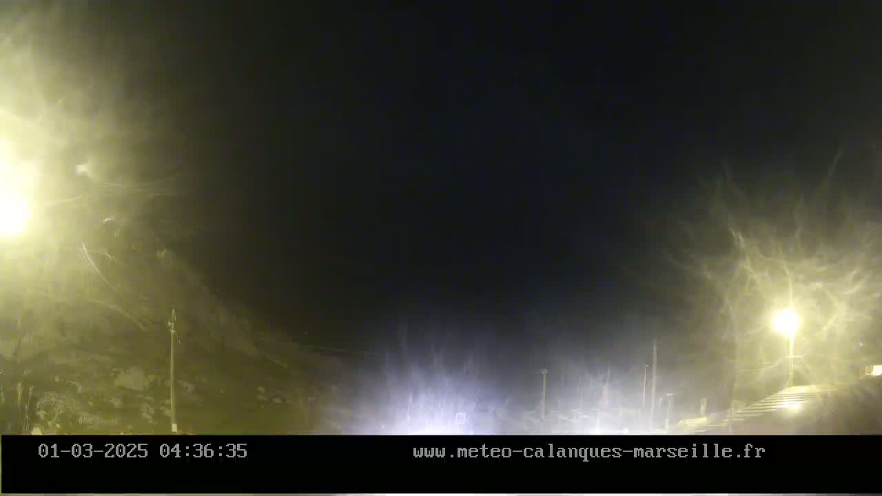 A nighttime outdoor scene shows blowing snow obscuring the view of a road and surrounding area illuminated by streetlights.