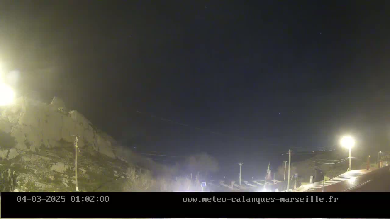 A nighttime view of a sparsely lit road leading to a cliff face under a clear, dark sky.