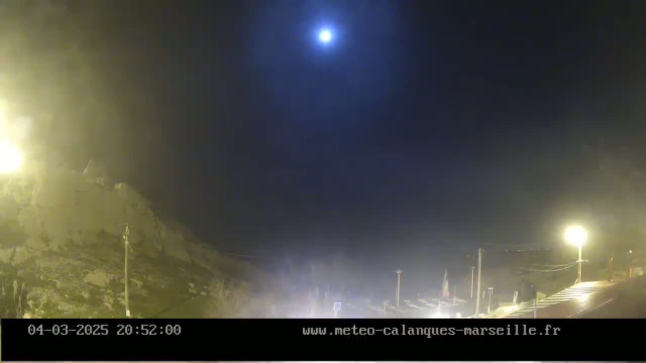 A bright moon illuminates a hazy, nighttime scene of a rocky hillside and a roadway with some parked cars and streetlights.