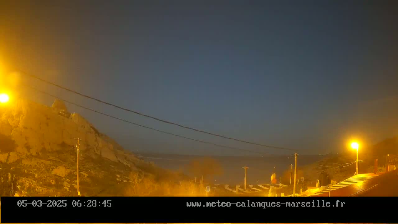 A dark, hazy twilight scene shows a rocky hill illuminated by streetlights overlooking a calm body of water.