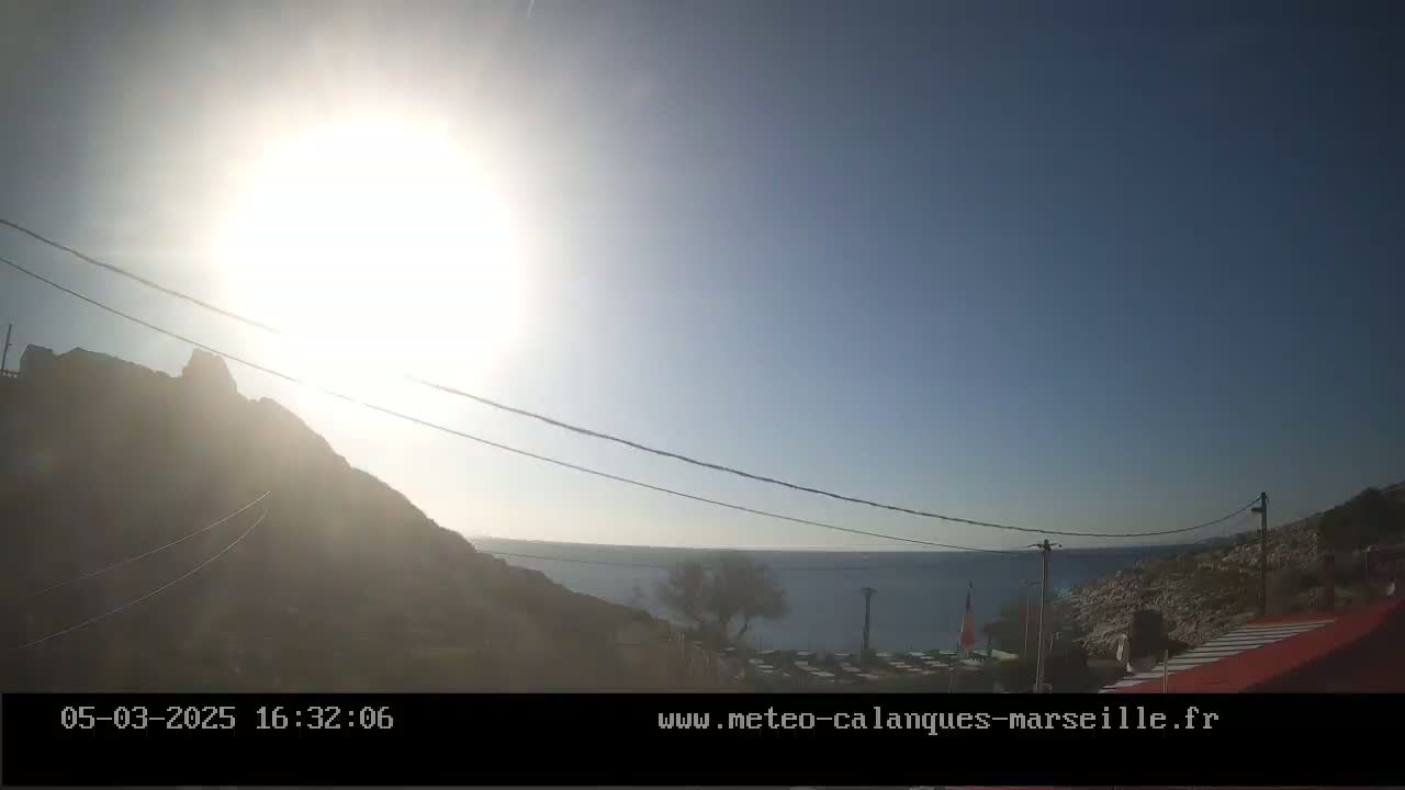 A bright, sunny day shows a rocky hillside overlooking a calm body of water, with power lines stretching across the scene.