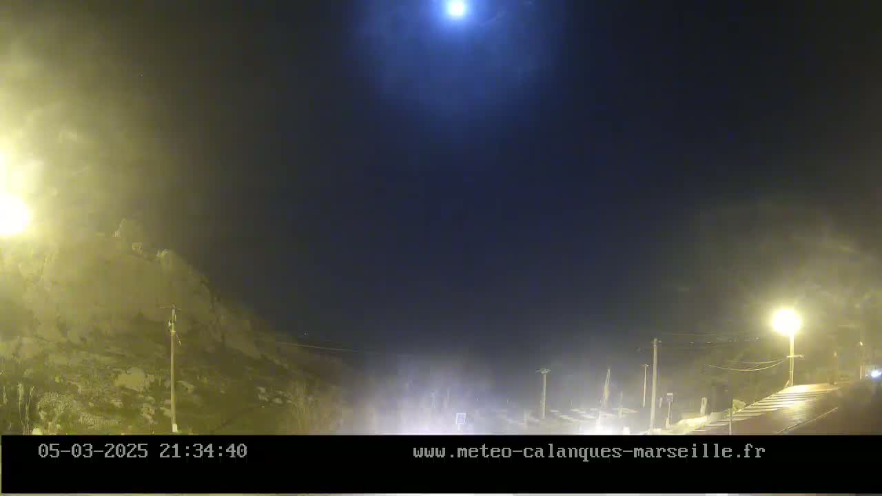 A nighttime view of a rocky hillside under a hazy, moonlit sky shows a road with some light traffic.