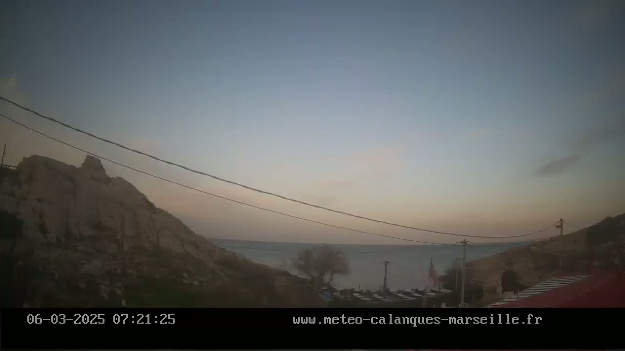 A calm, dusky sunrise over a calm body of water, viewed from a rocky hillside with power lines overhead.