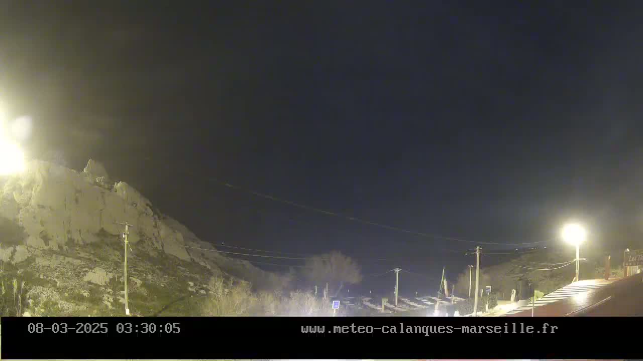 A nighttime view of a rocky hillside, sparsely vegetated, under a clear, dark sky, with a few streetlights illuminating a roadway and some parked vehicles in the foreground.