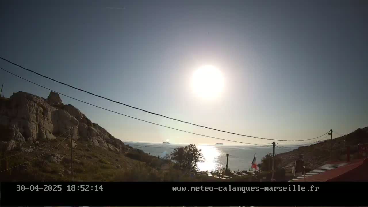 A bright sun shines over a calm sea, with rocky hillsides and power lines in the foreground.