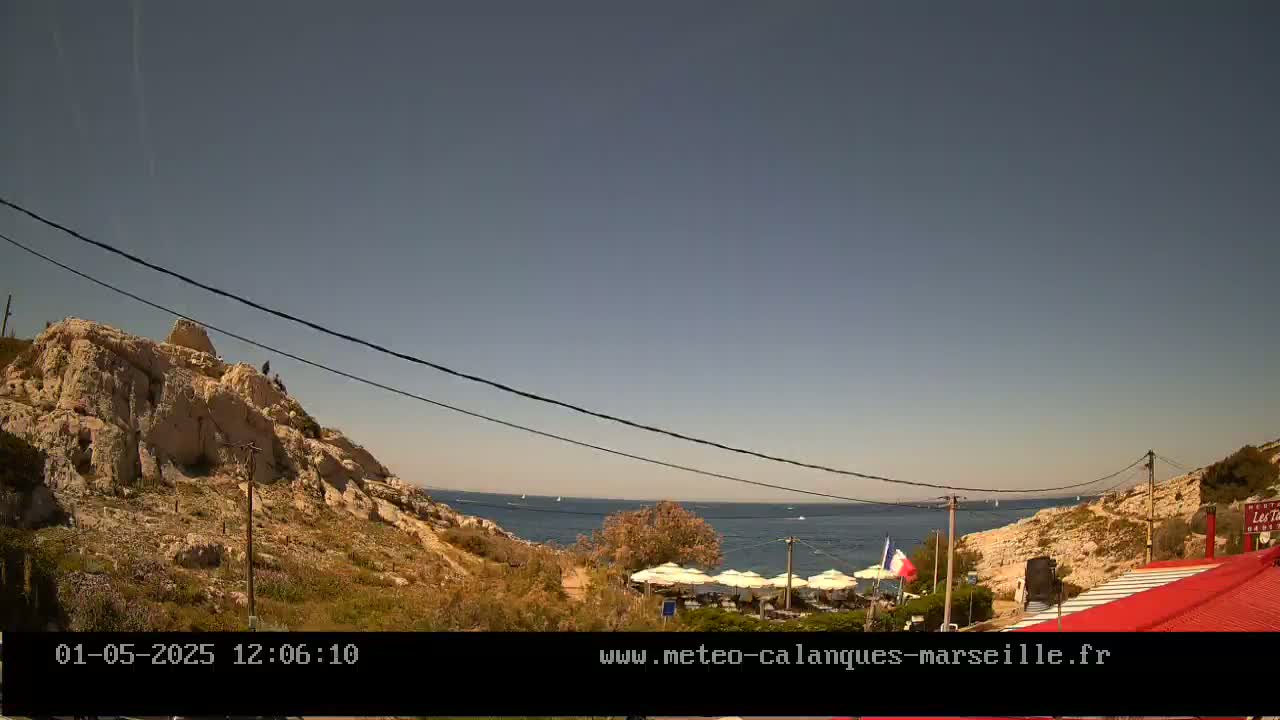A rocky hillside overlooks a calm sea with a few boats visible, under a clear, sunny sky; a restaurant with umbrellas is situated on the coast below.