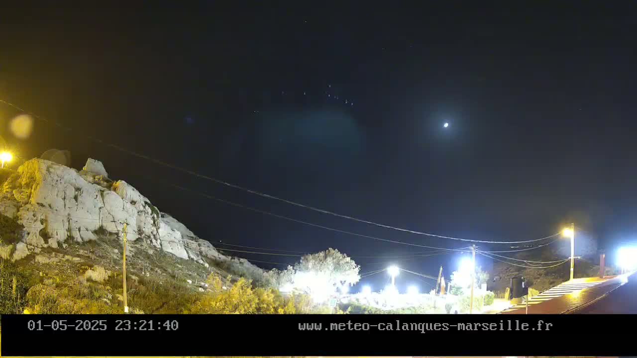 A nighttime view of a rocky hillside illuminated by streetlights under a clear sky with a visible moon and stars.