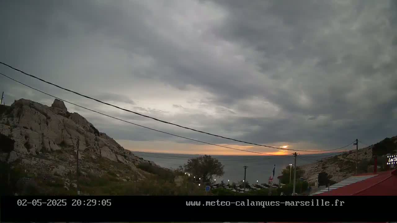 A rocky hillside overlooks a calm sea at sunset under a mostly cloudy sky.