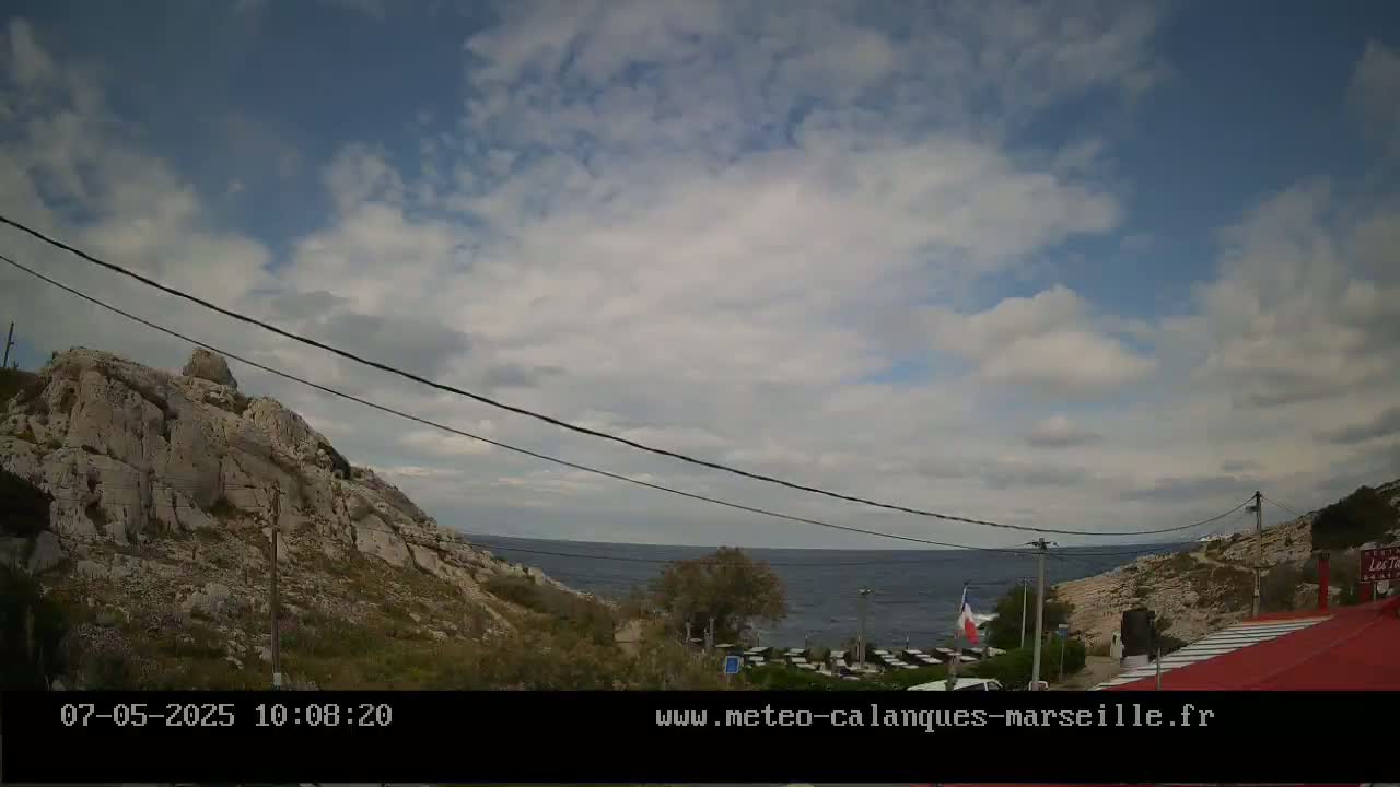 A partly cloudy sky overlooks a rocky coastline with a calm sea, a restaurant with a red roof, and power lines running across the scene.