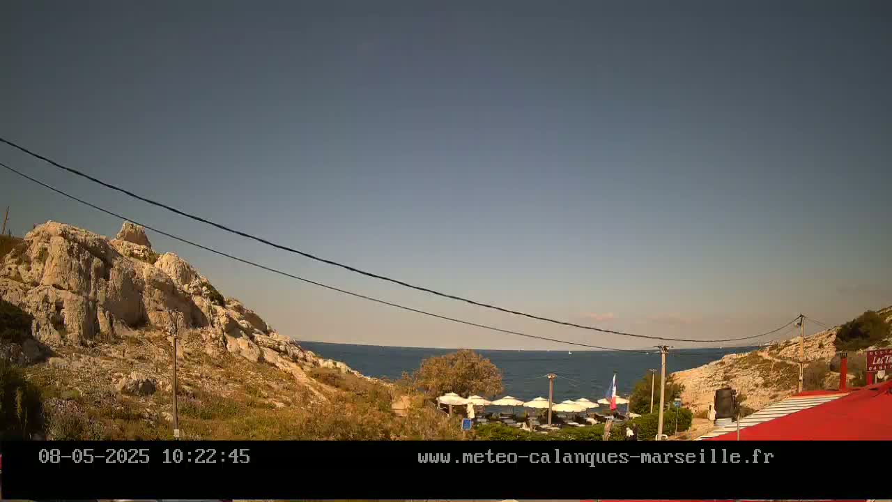 A rocky hillside overlooks a calm sea with a few boats visible in the distance under a clear, sunny sky; a restaurant with patio umbrellas is situated near the water's edge.