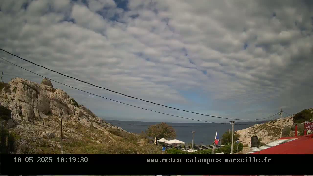 A partly cloudy sky overlooks a rocky coastline with a small restaurant and beach umbrellas visible in the distance.