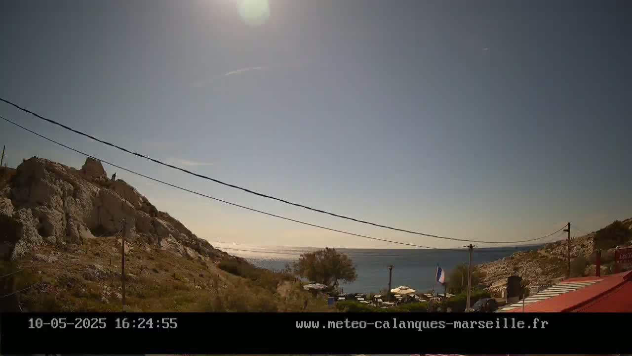A sunny day overlooking a calm sea from a rocky hillside with sparse vegetation, featuring a small outdoor seating area.