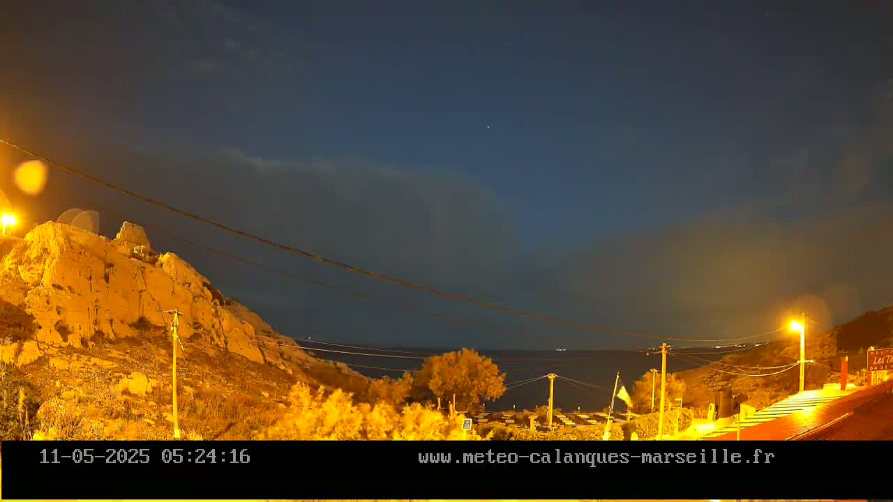 A nighttime coastal scene shows a rocky hill illuminated by artificial light, overlooking a calm sea under a mostly clear, dark sky.