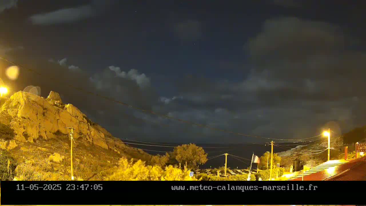 A nighttime view of a rocky hillside overlooking a calm sea under a partly cloudy sky.