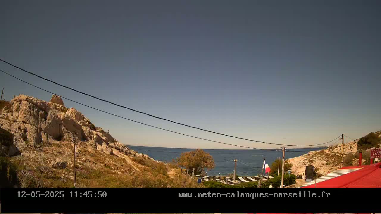 A clear, sunny day overlooks a rocky coastline with a calm sea and a glimpse of outdoor seating at a restaurant.