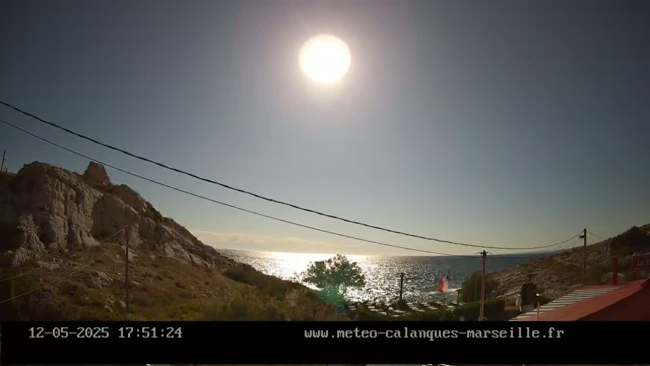 A bright sun shines over a rocky shoreline and calm sea, with power lines stretching across the view.