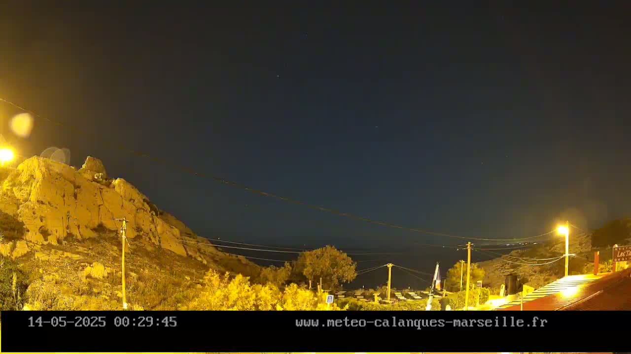 A nighttime view shows a rocky hillside illuminated by artificial lights, overlooking a calm body of water under a clear, dark sky.