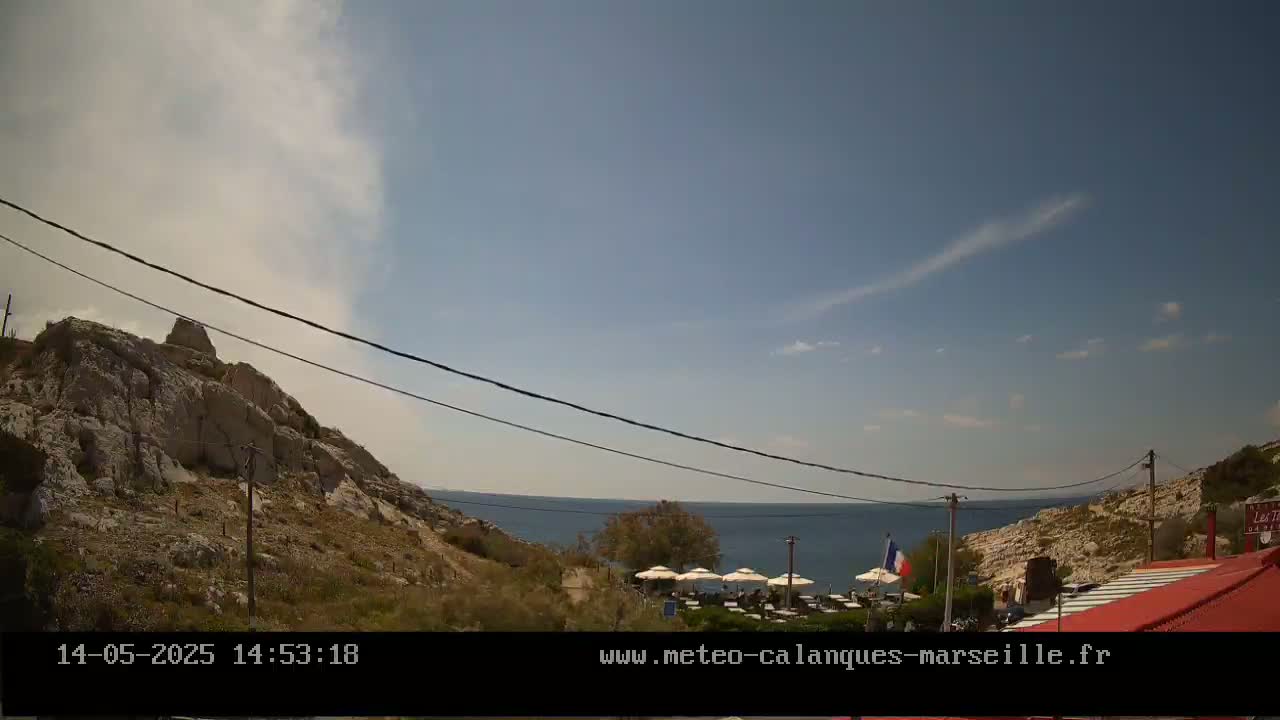 A rocky hillside overlooking a calm sea, under a mostly clear sky with a hazy plume of smoke or cloud in the distance, features a restaurant with patrons visible in the foreground.