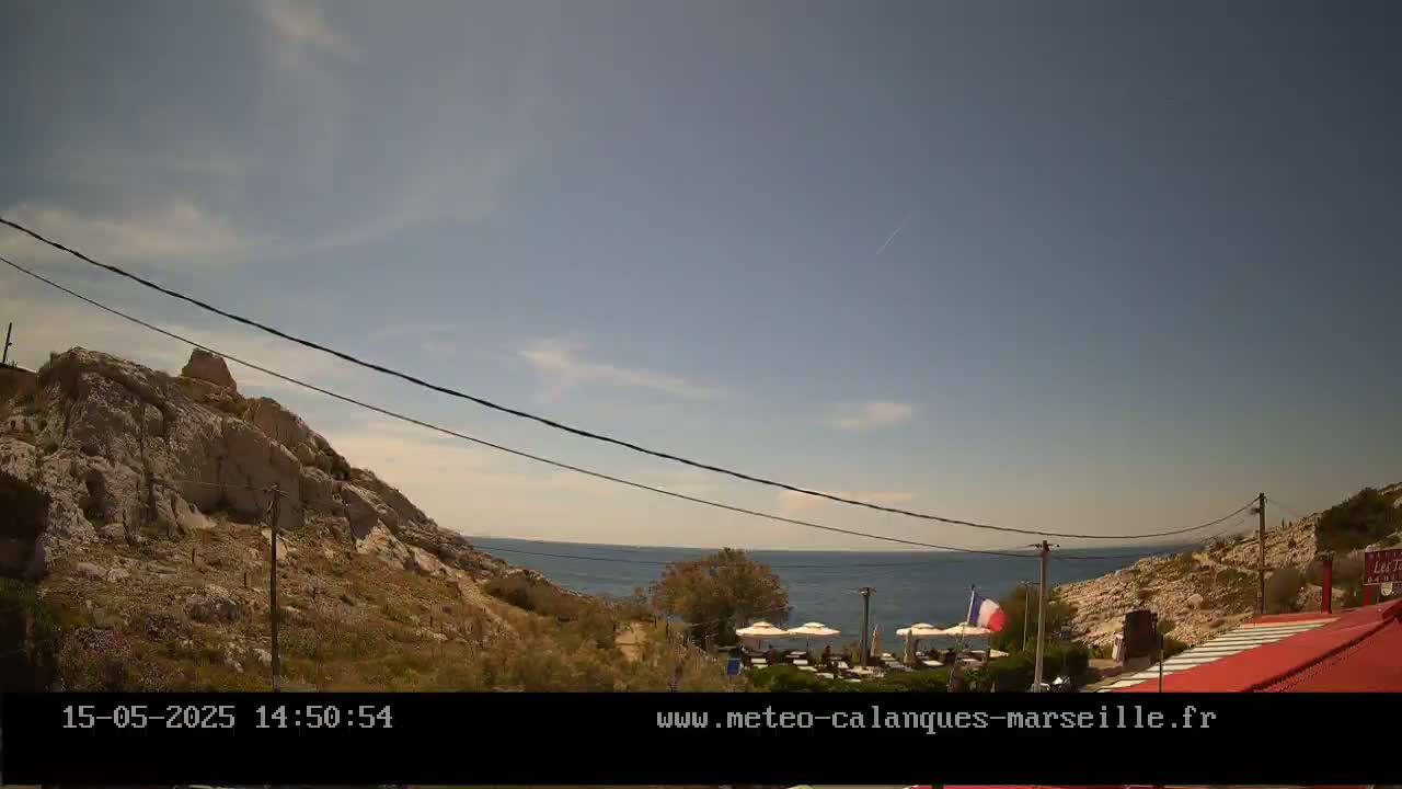 A mostly sunny day shows a rocky, Mediterranean coastline with a restaurant and a French flag visible near the water.