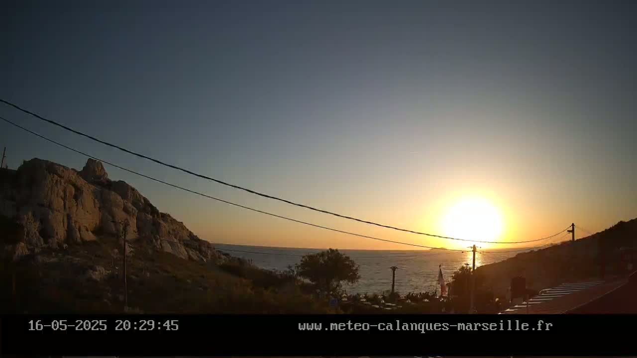 A sunset over a calm sea, with a rocky hillside in the foreground and power lines crossing the frame.