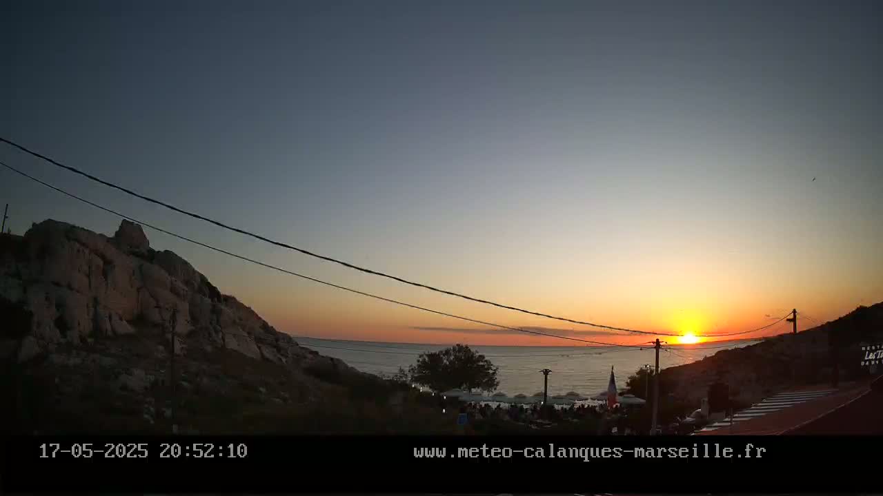 A sunset over a calm sea, viewed from a rocky hillside with a gathering of people near the water's edge.