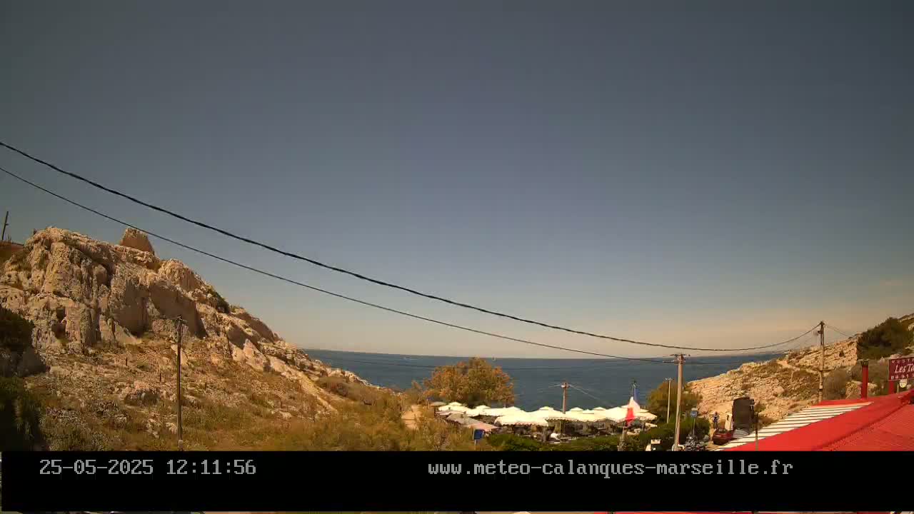 A sunny day reveals a rocky coastline with a calm sea, a small restaurant with outdoor seating, and power lines strung overhead.