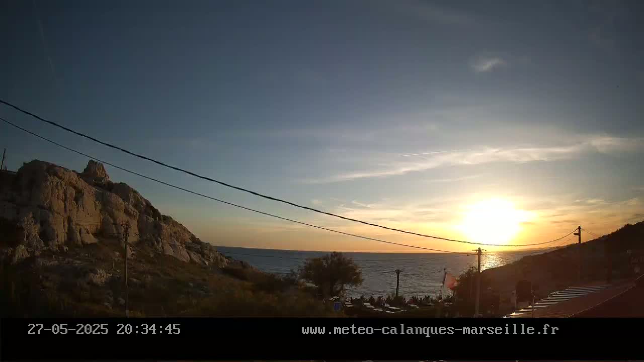 A sunset over a calm sea, with a rocky hill to the left and a few people gathered near the water's edge.