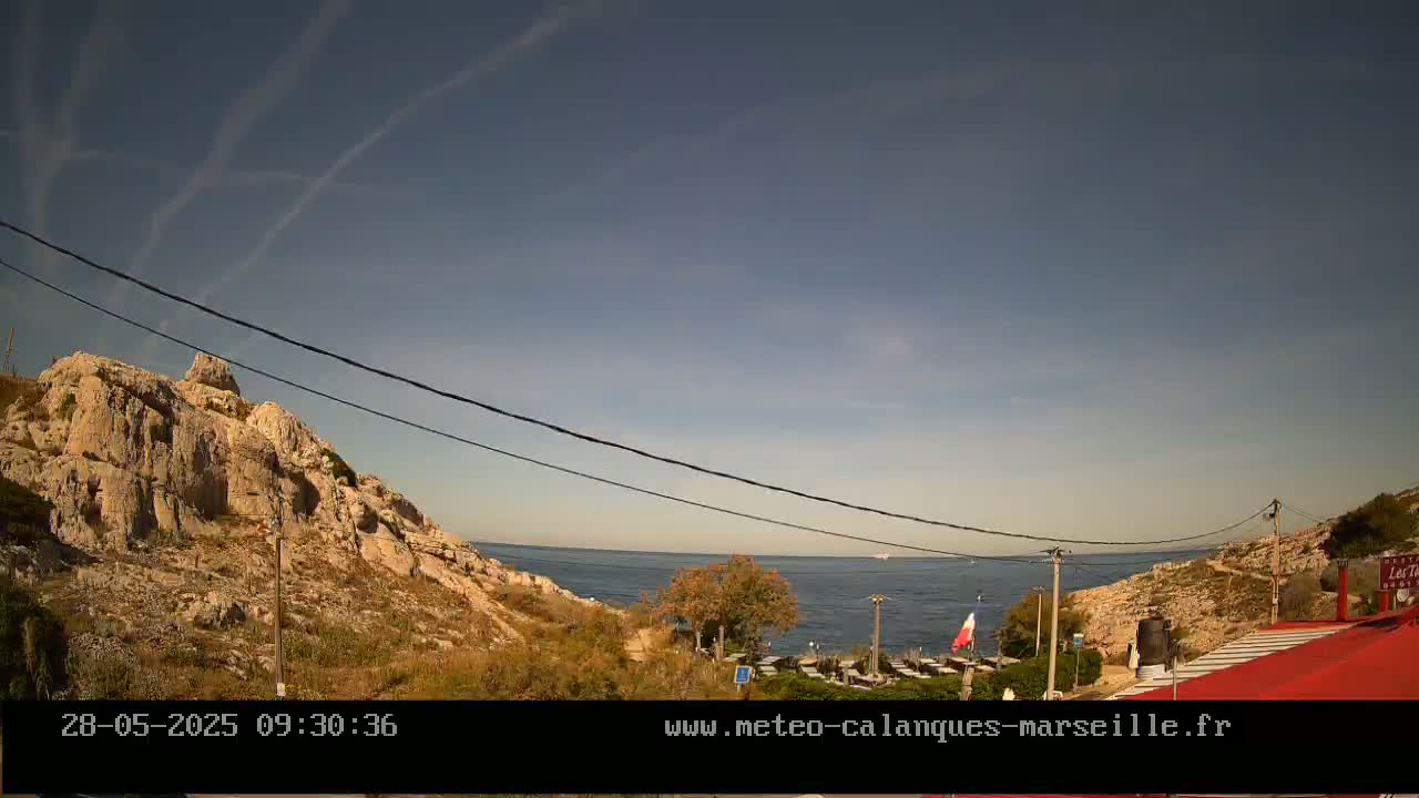 A mostly clear sky over a rocky coastline with a calm sea, a small beach, and some outdoor seating at a restaurant.