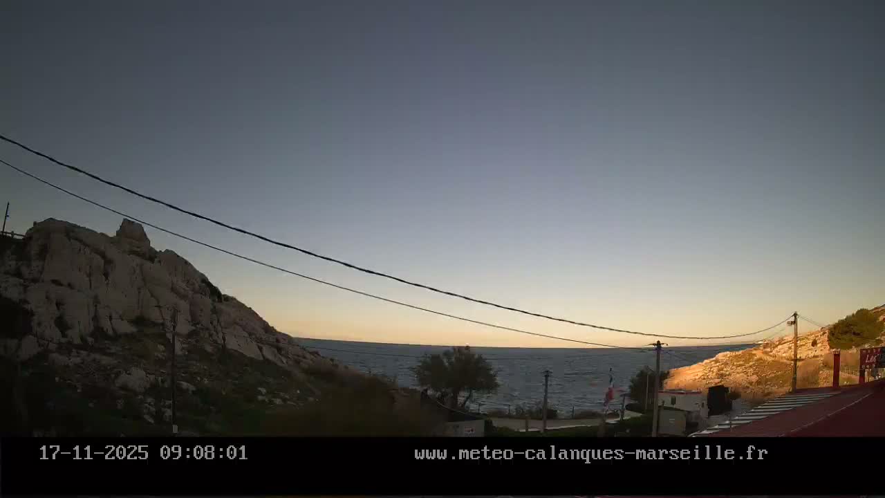 A tranquil coastal landscape under a clear, gradient sky transitioning from deep blue to warm yellow at the horizon, features calm blue waters framed by rocky hills with sparse vegetation, on a fair weather day.