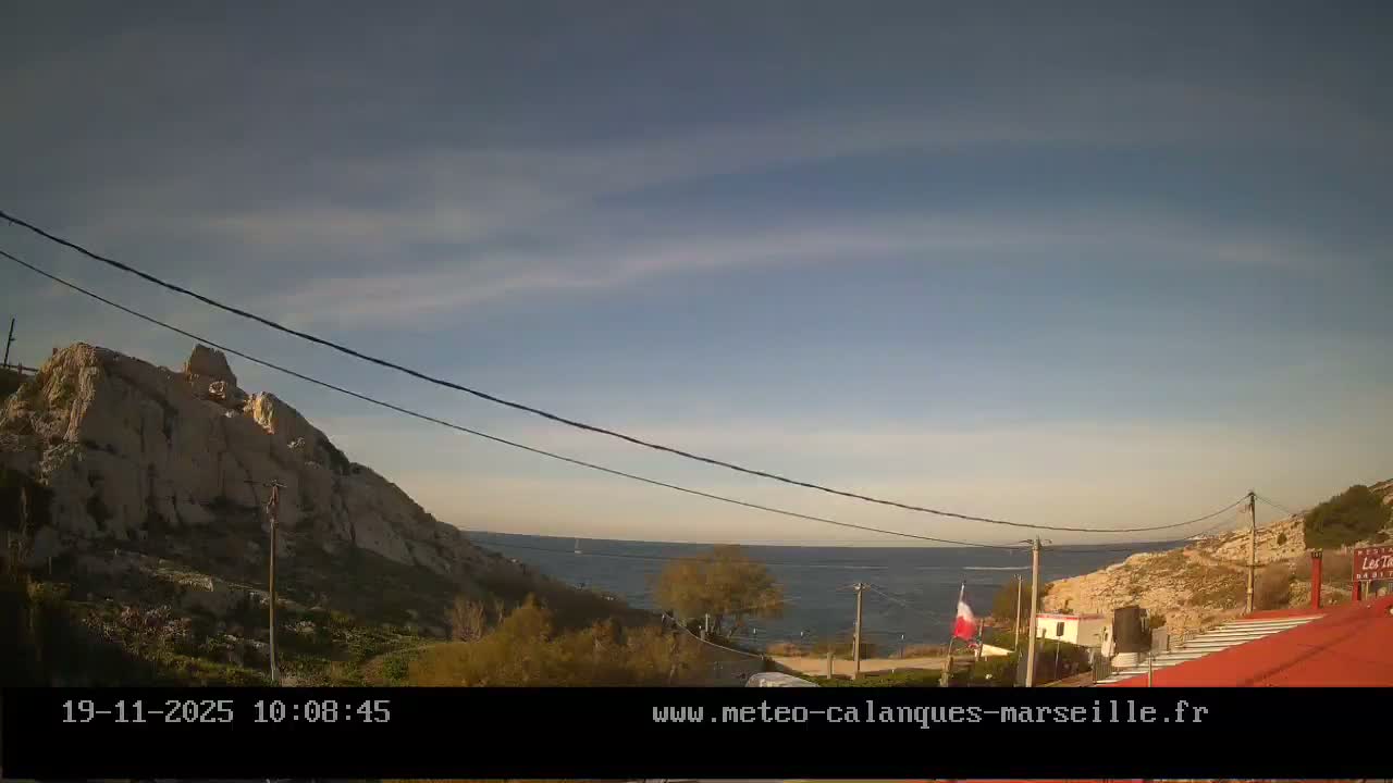 The image displays a bright coastal landscape featuring rugged rocky mountains, a calm blue sea with distant boats, and red-roofed buildings nestled on a hillside, all under a clear to partly cloudy sky.