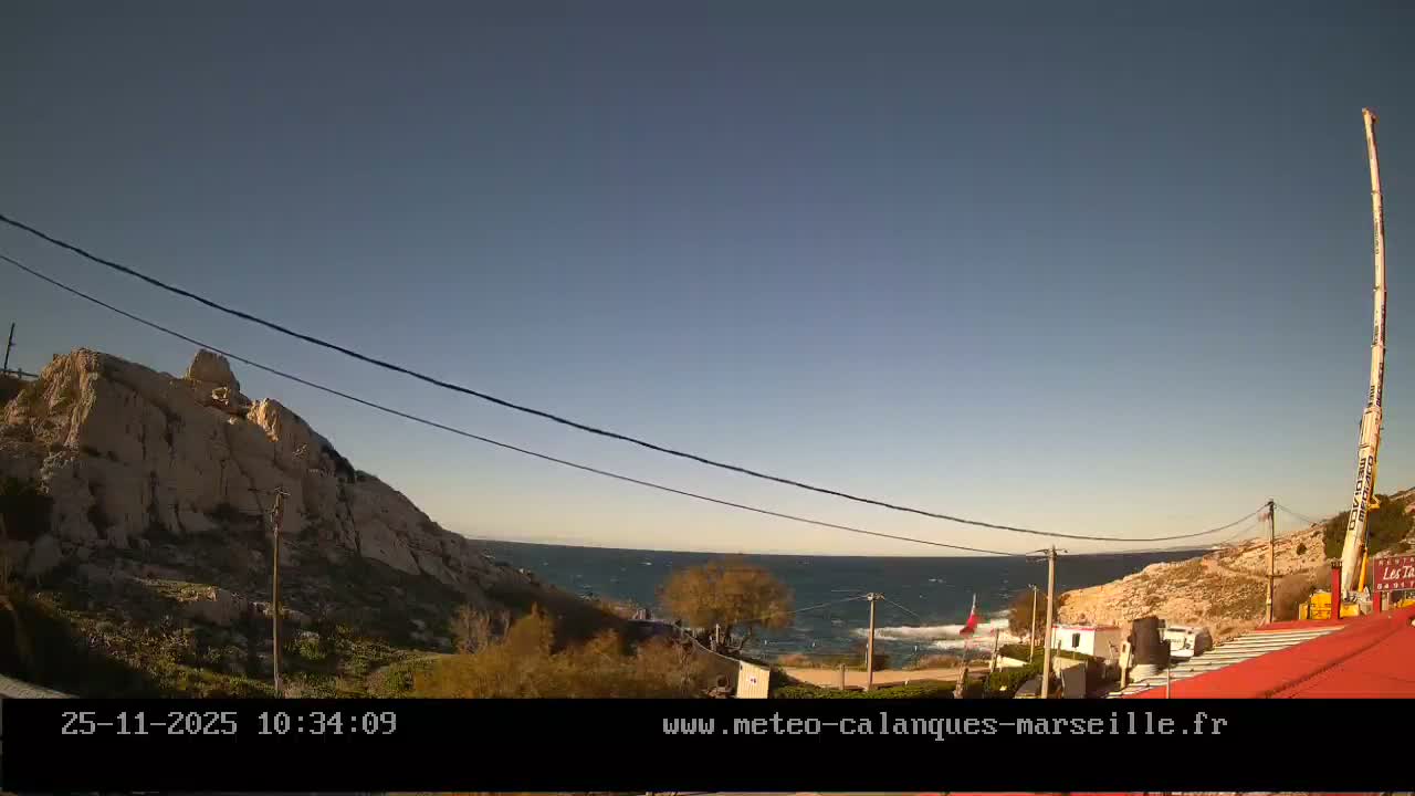 The image displays a clear, sunny day over a rugged coastal landscape with a calm blue sea, rocky hills, power lines strung across the frame, and a tall white crane on the far right.