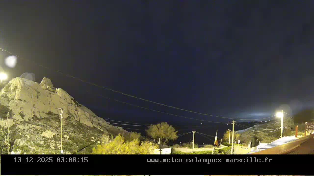 A rocky, pale cliff dominates the left side of a nighttime landscape under an overcast sky, with scattered artificial lights illuminating sparse vegetation and a path on the right.