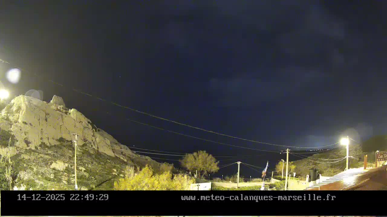 A rocky, pale cliff dominates the left side of a nighttime landscape under an overcast sky, with scattered artificial lights illuminating sparse vegetation and a path on the right.
