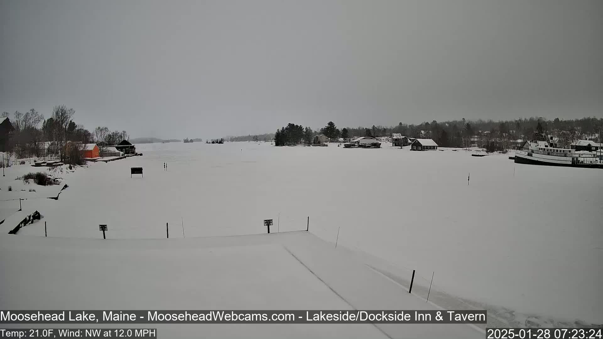 A snow-covered frozen lake is seen from a lakeside viewpoint, with some buildings and a boat visible in the distance under an overcast sky.