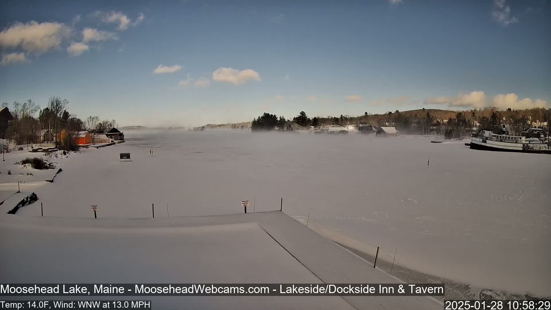 A snow-covered frozen lake with a light fog, buildings on the shore, and a boat in the distance under a partly cloudy sky.