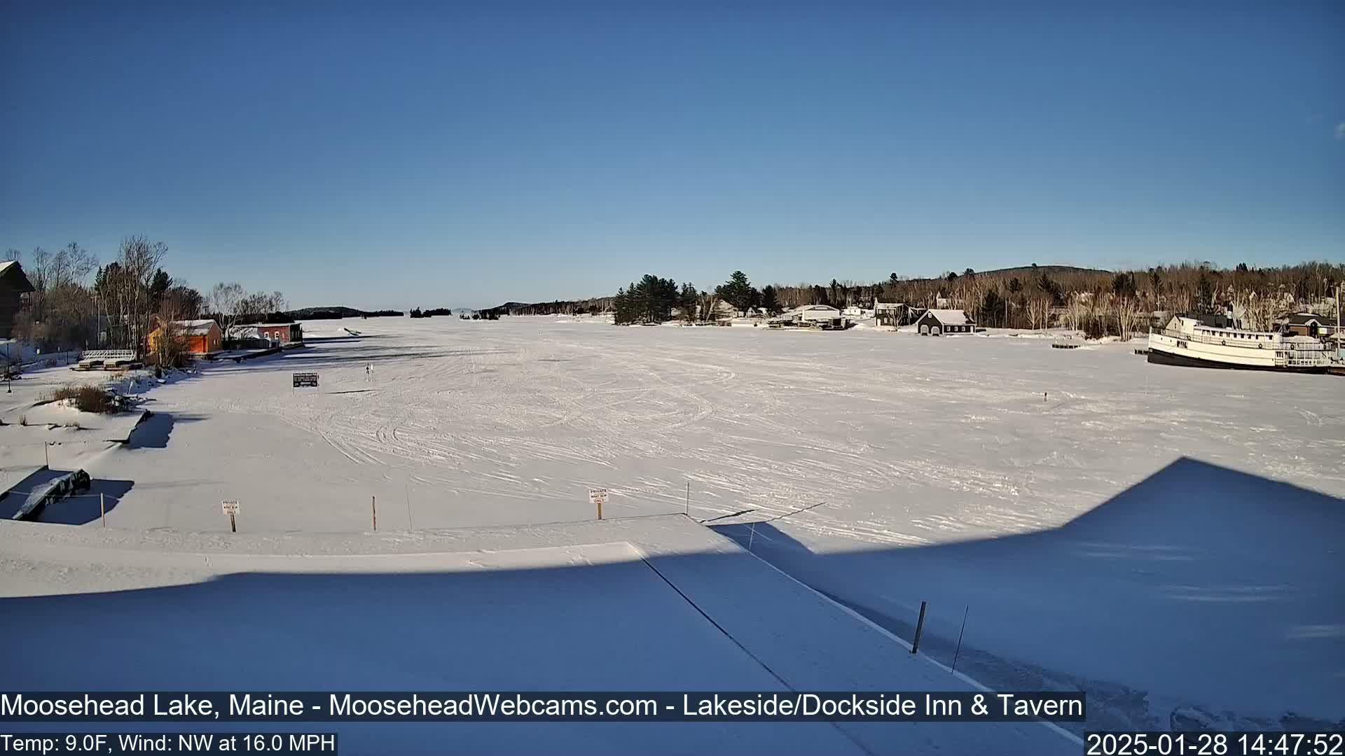 A frozen lake under a clear blue sky, with a few buildings and a boat visible on the shore, and a strong northwest wind.