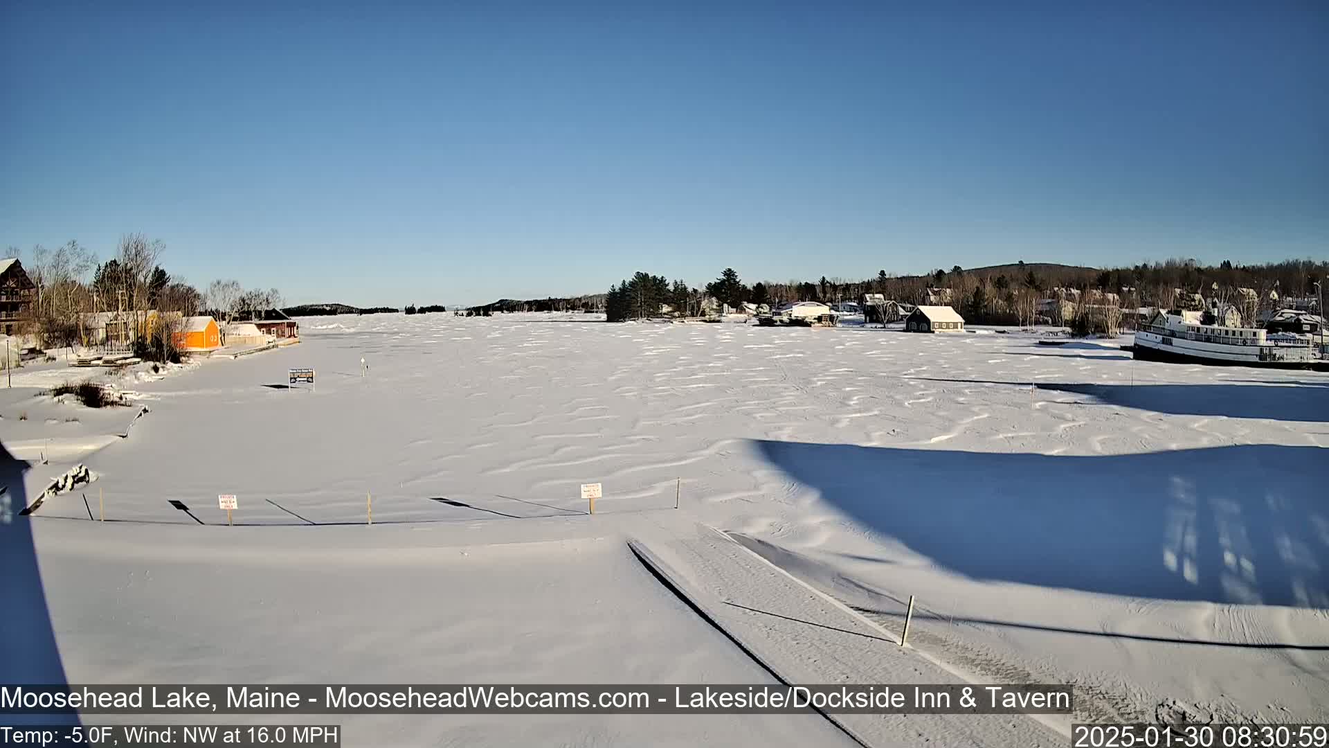 A snow-covered frozen lake stretches out from a shoreline with buildings and a docked boat under a clear blue sky.