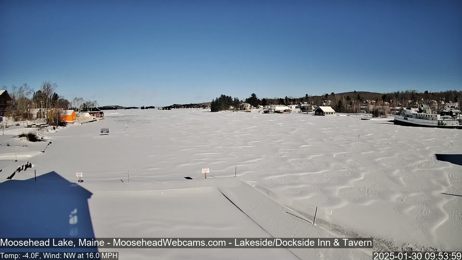 A snow-covered frozen lake with some buildings and a boat visible in the distance under a clear blue sky, with a northwest wind at 16.0 mph and a temperature of -4.0°F.