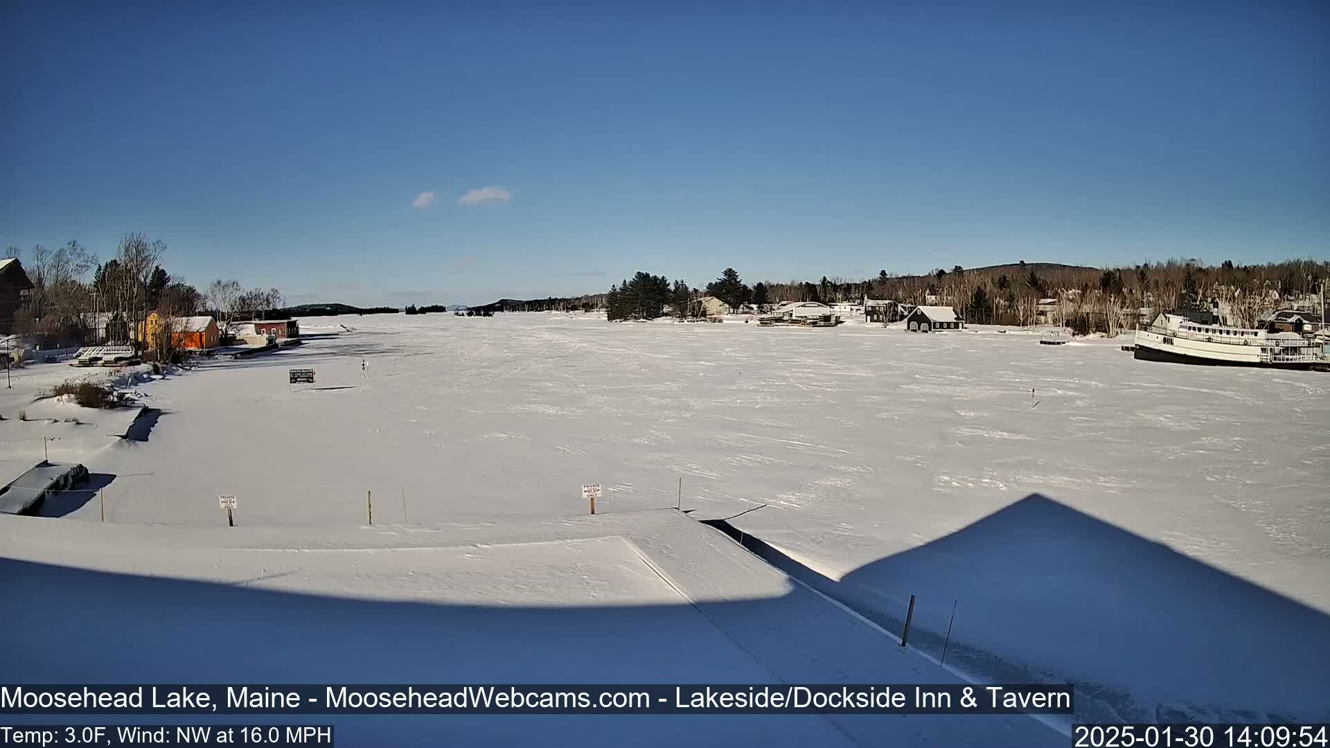 A frozen lake stretches out under a clear blue sky, showing a few buildings and a boat on the snow-covered surface.