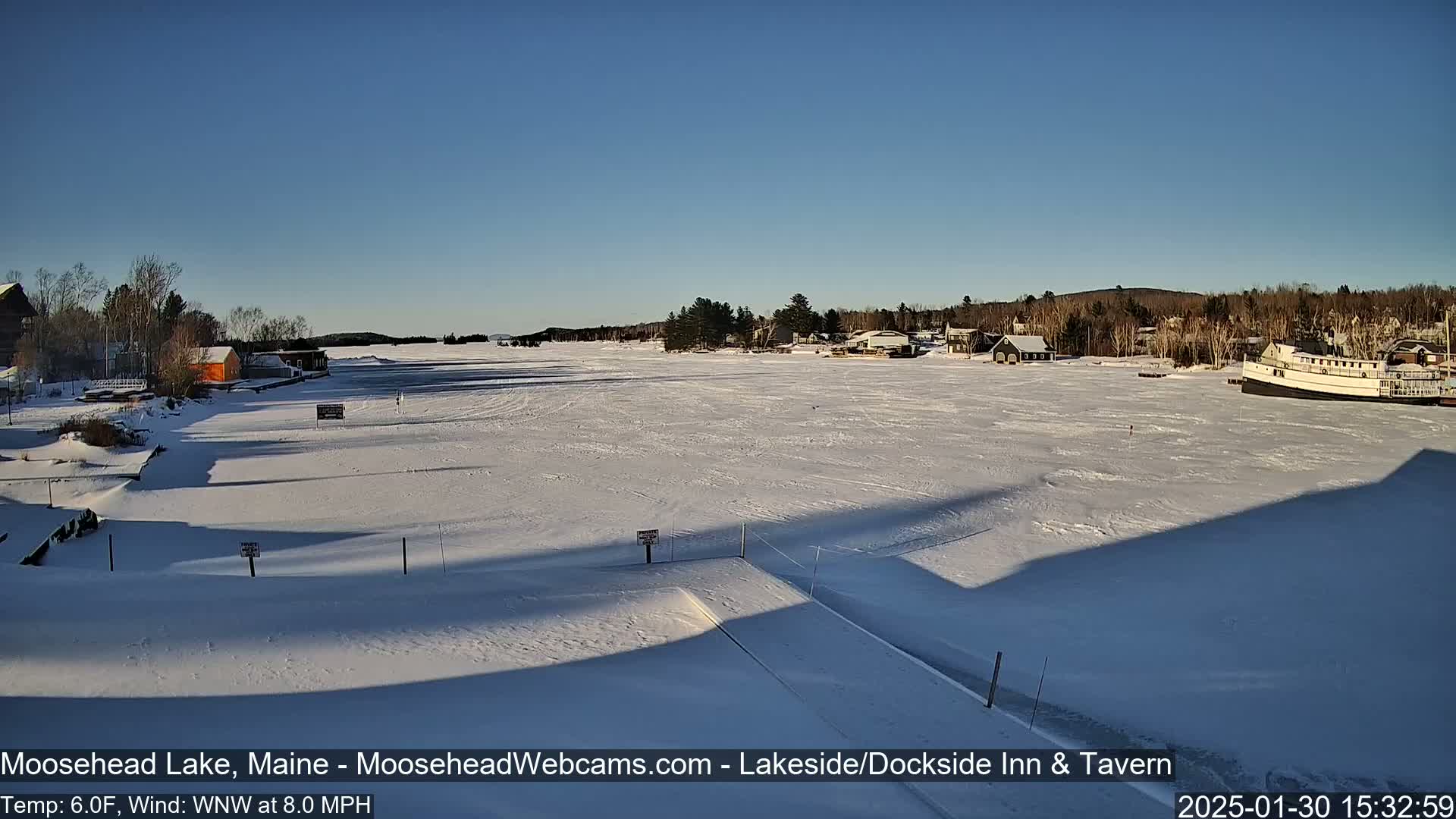 A frozen lake, under a clear blue sky, is seen from a snowy vantage point, with several buildings and a boat visible on the shore.